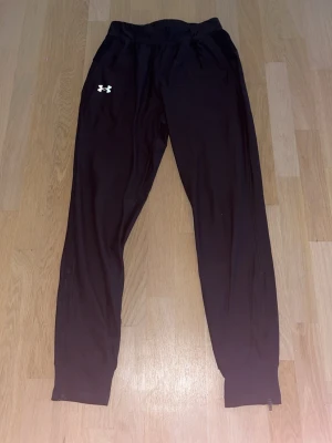 Svarta träningsbyxor Under Armour - Säljer ett par svarta träningsbyxor från Under Armour med smal passform och dragkedjor vid bensluten. Byxorna har resår i midjan och logga på vänster lår. Perfekta för gymmet eller löpning.
