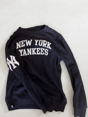  New York Yankees sweatshirt - Cool svart sweatshirt med vit 'NEW YORK YANKEES'-text på bröstet och Yankees-logga på ärmen. Tröjan har rund halsringning och långa ärmar. Perfekt för dig som gillar sportig streetstyle och vill representera ditt favoritlag.