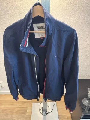 Marinblå vindjacka från Hilfiger Denim - Snygg marinblå vindjacka från Hilfiger Denim med dragkedja och ribbade muddar med röd, vit och blå rand. Jackan har ståkrage och diskret logga på bröstet. Perfekt för dig som vill ha en stilren och sportig look. Materialet är lätt och vindtätt.