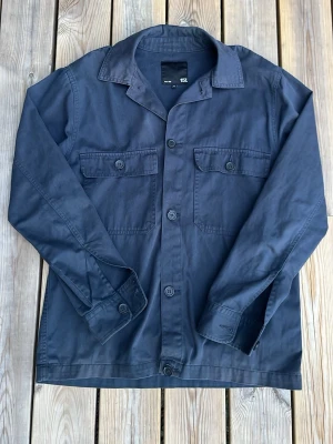 Mörkblå overshirt med fickor - Stilren mörkblå overshirt med två bröstfickor med lock och knappar. Klassisk krage och knäppning framtill. Tillverkad i ett mjukt bomullstyg som ger en avslappnad look. Perfekt lager på lager-plagg för dig som gillar minimalistisk stil.