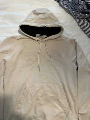 Beige hoodie från Burberry - Säljer en beige hoodie från Burberry med broderad logga på bröstet. Hoodien har en stor ficka framtill, dragsko i huvan och svart innerfoder i huvan. Tröjan är slimfit och skulle passa perfekt för den med storleken xs-s
