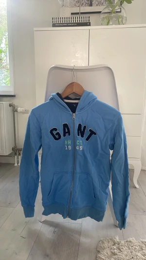 Blå zip hoodie från GANT - Snygg ljusblå hoodie från GANT med dragkedja framtill och broderad logga på bröstet. Tröjan har huva, två fickor och ribbade muddar vid ärmar och nederkant. Perfekt för en avslappnad och sportig stil.