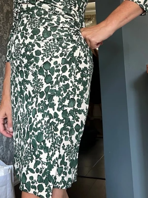 Tory Burch Grönvit omlottklänning med bladmönster - Snygg omlottklänning från trendiga Tory Burch med grönt bladmönster på vit botten. Klänningen har trekvartsärm, v-ringning och rynkade detaljer i sidan som ger en smickrande passform. Tyget är tätt och silkigt. Perfekt för dig som gillar naturinspirerade prints och vill ha något bekvämt men ändå stilrent.