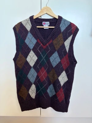 Vintage stickad väst - Royal Scott - Vintage väst med argylemönster. Begagnad men gott skick. Finns ett hål på ena sidan där blått garn trätts igenom för att laga det. Detta är inget som märks då det är på ett område utom synhåll. Vill man fixa hålet själv med rätt teknik finns möjligheten då insidan inte är ”säkrad”. Garnet hänger alltså bara och lättare att dra loss för att ersätta med nytt. Vill man ha det som det är går det också. Material: ull. Följ tvättanvisningarna. Har katt hemma. Köpt för 716kr.