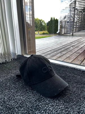 ALO Yoga keps - Säljer en svart keps med böjd skärm och justerbart spänne bak. Framtill finns en diskret broderad logga i svart ton-i-ton. Klassisk och stilren modell som passar till det mesta. Materialet ser ut att vara bomull.