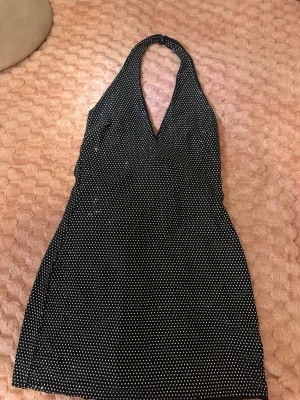 Svart glittrig halterneckklänning - Snygg svart kort klänning med glittriga detaljer och halterneck. Klänningen har v-ringning och är ärmlös, perfekt för en partylook. Materialet känns mjukt och följsamt, och den har ett diskret prickigt mönster av glitter.