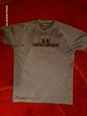 Grå träningstopp från Under Armour - Snygg grå träningstopp från Under Armour med korta ärmar och rund hals. Toppen har logga och text framtill och är tillverkad i ett lätt och funktionellt syntetmaterial som passar perfekt för aktiva dagar.