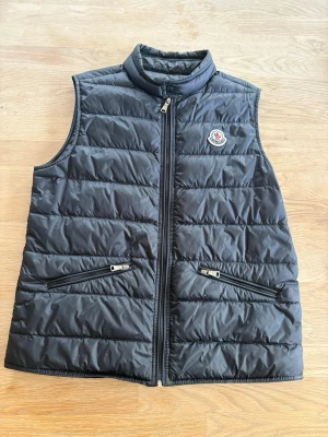 Svart dunväst från Moncler - Snygg svart dunväst från Moncler med quiltad design och hög krage. Västen har två dragkedjeförsedda fickor framtill och Moncler-logga på bröstet. Perfekt för lager-på-lager och håller dig varm med lätt dunfyllning.