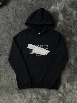 Svart Maison Margiela hoodie - Säljer en svart hoodie från Maison Margiela med tryck framtill och klassisk logga. Hoodien har huva, magficka och ribbade muddar. Tillverkad i mjukt bomullsmaterial och har en avslappnad passform. Perfekt för dig som gillar streetwear och unik design.