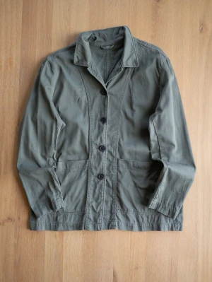 Olivgrön overshirt med knappar - Snygg olivgrön overshirt i bomull med klassisk krage och stora svarta knappar framtill. Jackan har två stora fickor framtill och raka ärmar. Perfekt för lager på lager och enkel att styla till olika outfits.