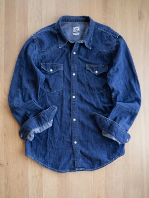 Mörkblå jeansskjorta från Lee - Säljer en klassisk mörkblå jeansskjorta från Lee med tryckknappar och två bröstfickor. Skjortan har lång ärm och kontrastsömmar som ger en snygg vintagekänsla. Perfekt till jeans eller chinos för en avslappnad look.