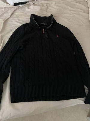 Svart kabelstickad tröja Polo Ralph Lauren - Kabelstickad svart tröja från Polo Ralph Lauren med halv dragkedja och hög krage. Tröjan har ett rött broderat logomärke på bröstet och ribbade muddar. Perfekt för dig som gillar klassisk och stilren design. Det är en storlek M men passar oxå för den som har storkek S 