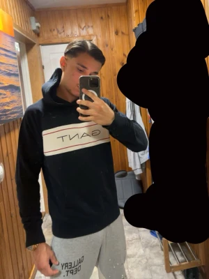 Svart hoodie från GANT med logga - Säljer en svart hoodie från GANT med stor vit logga och röda linjer över bröstet. Klassisk huva och magficka, ribbade muddar vid ärmslut och nederkant. Perfekt för en avslappnad och stilren look.