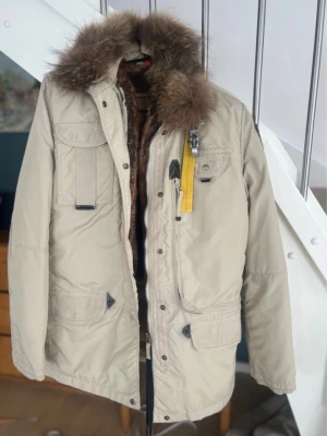Beige parkas med  från Parajumpers - Stilren beige parkas från Parajumpers med avtagbar huva och brun fuskpäls på insidan och runt kragen. Jackan har flera praktiska fickor, gul detalj vid dragkedjan och robust dragkedja. Perfekt för kalla dagar och riktigt snygg till vintern. Liten i storlek som alla deras kläder