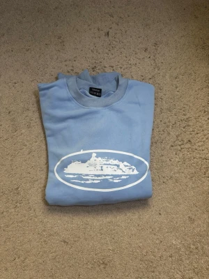 Ljusblå sweatshirt med fartygstryck från Corteiz - Ljusblå sweatshirt med rund hals och ribbade muddar vid ärmar och nederkant. På bröstet finns ett vitt tryck av ett fartyg inuti en oval. Perfekt för dig som gillar marint tema och avslappnad stil.