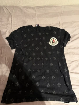 Svart Moncler t-shirt med logga - Snygg svart t-shirt från Moncler med korta ärmar och rund hals. T-shirten har ett diskret mönster med små vita Moncler-loggor över hela plagget och en större logga på bröstet. Perfekt för dig som gillar streetwear och vill ha något stilrent men ändå med attityd.