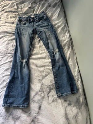 Blå bootcut jeans med slitningar - Säljer ett par blå bootcut jeans med slitningar på båda knäna och snygga röda detaljer på bakfickan. Jeansen har klassisk femficksdesign och är gjorda i denim med lätt tvättad look. Perfekta för en avslappnad och trendig stil.