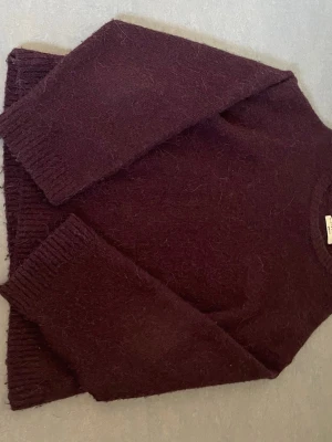 Mörklila stickad tröja - Mysig stickad tröja i mörklila färg med rund halsringning och långa ärmar. Tröjan har ribbstickade muddar vid ärmslut och nederkant. Materialet ser ut att vara mjukt och fluffigt, perfekt för kyliga dagar.