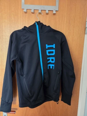 Svart hoodie med blå dragkedja och IDRE-tryck - Säljer en svart hoodie med blå dragkedja och stort blått IDRE-tryck på framsidan. Hoodien har huva, två fickor med dragkedja och ett vitt renmärke på ärmen. Materialet känns mjukt och stretchigt, perfekt för chill dagar.