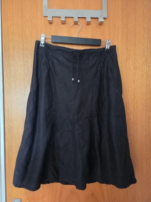 Svart långkjol med snörning - En svart långkjol i mjukt tyg med snörning i midjan och diskret A-linjeform. Kjolen har en enkel design utan mönster och dragkedja bak för enkel påklädning. Perfekt för dig som gillar stilrena plagg med en avslappnad vibe.