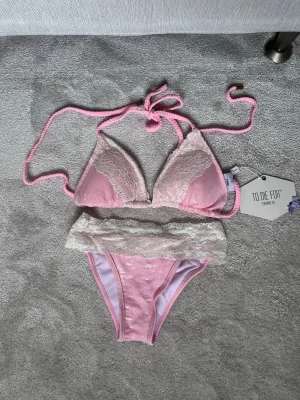 Rosa paljettbikini med spetsdetaljer - Supersöt rosa paljettbikini från To Die For med vita spetsdetaljer på både överdel och underdel. Bikinin har knyt i nacken och ryggen samt små glittriga paljetter på tyget. Perfekt för sommarens strandhäng!