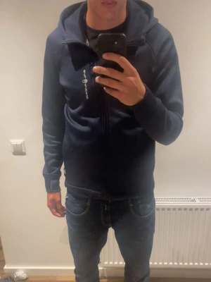 Mörkblå hoodie från Sail Racing - Snygg mörkblå hoodie från Sail Racing med dragkedja framtill och två sidofickor med dragkedja. Märkets logga syns på bröstet och huvan har en liten logodetalj. Tillverkad i mjukt material, perfekt för chill eller uteaktiviteter.