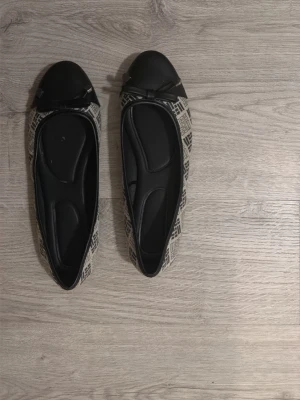 Svarta ballerinaskor med mönster - Snygga ballerinaskor med svart tåparti och rosett framtill. Skorna har ett beige tyg med svart grafiskt mönster längs sidorna och svarta detaljer. Klassisk rundad tå och låg sula. Perfekta för dig som gillar stilrena men ändå unika skor.