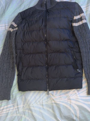 Moncler cardigan Grå - Snygg cardiganjacka från Moncler med vadderad  front och kabelstickade grå ärmar. Ärmarna har vita ränder och Moncler-logga på vänster sida. Jackan har dragkedja framtill och två fickor med dragkedja. Perfekt för lager-på-lager och streetstyle. Tyvärr så finns några små fläckar på framsidan som man ser på bilden men utöver det så ingen ting annat, och Scan fungerar med som sista bilden visar och för mer frågor och funderingar så kan man skriva i pm MVH!