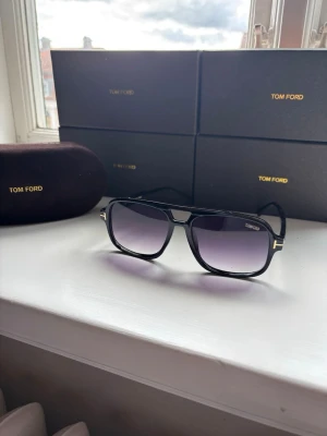 Svarta Tom Ford solglasögon - Snygga svarta solglasögon från Tom Ford med fyrkantig form och lila tonade glas. Bågarna har guldfärgade detaljer vid sidorna och en klassisk, stilren look. Perfekta för att lyfta din outfit under soliga dagar.