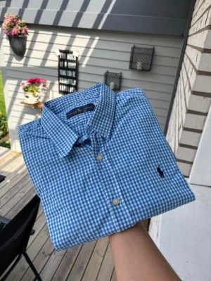 Blårutig skjorta från Ralph Lauren - Säljer en klassisk blårutig skjorta från Ralph Lauren med button-down krage och lång ärm. Skjortan har vita knappar framtill och den ikoniska broderade loggan på bröstet. Perfekt till jeans eller chinos för en clean och stilren look.