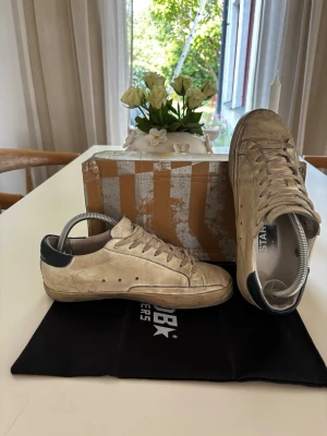 Golden goose skor - Snygga beige sneakers från Golden Goose med svart stjärndetalj på sidan och svart hälparti. Skorna har klassisk låg modell, vita snören och slitna detaljer för en cool, avslappnad look. Perfekta för dig som gillar streetstyle och unika detaljer. Storlek 41, använda hyfsat mycket, men varsamt. Allt på bilden tilkommer, dustbag och box. Med en liten tvätt blir dom som nya! 🛀🫧