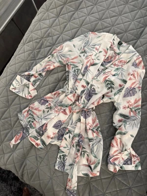 Blommig kimono-blus med knytband - Snygg blus i kimonomodell med blommigt mönster i pastellfärger på vit botten. Har långa ärmar och ett matchande knytband i midjan. Perfekt för en chill och trendig look. Materialet känns lätt och luftigt, troligen polyester eller viskos.