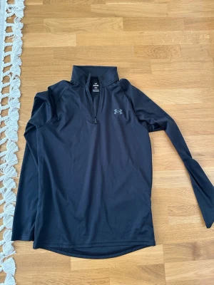 Svart träningströja från Under Armour - Svart långärmad träningströja från Under Armour med half zip och hög krage. Perfekt för löpning eller gym. Tillverkad i ett mjukt och stretchigt syntetmaterial som andas bra. Diskret logga på bröstet.