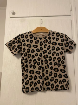 Leopardmönstrad t-shirt från Lindex - Säljer en beige t-shirt från Lindex med coolt leopardmönster i svart och brunt. T-shirten har korta ärmar och rund halsringning. Perfekt för dig som vill sticka ut lite extra i vardagen.