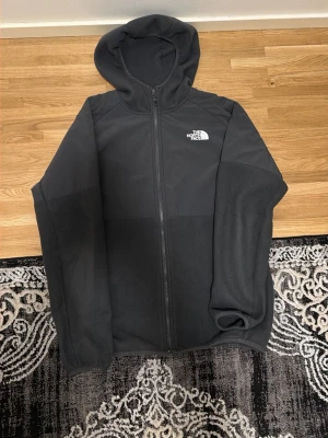 Grå fleecehoodie The North Face - Mysig grå fleecehoodie från The North Face med dragkedja framtill och huva. Tröjan har logga på bröstet och ryggen, samt långa ärmar. Perfekt för kyliga dagar och enkel att matcha med andra plagg. Storlek M