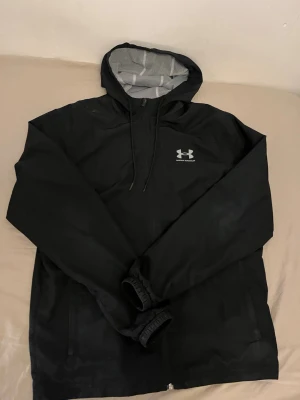 Svart vindjacka från Under Armour - Snygg svart vindjacka från Under Armour med huva och meshfoder. Jackan har dragkedja upptill, justerbar huva med snören och elastiska muddar vid ärmsluten. Perfekt för blåsiga dagar och sportiga outfits.