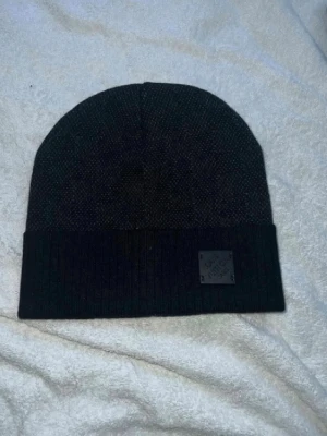 Svart ribbad mössa med patch - Snygg svart mössa med ribbad kant och en liten patch framtill. Klassisk modell som passar till det mesta och håller dig varm under kalla dagar. Materialet ser ut att vara stickat och mössan har en enkel, stilren look.
