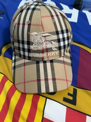 Beige Burberry keps med rutigt mönster - Säljer en klassisk beige keps från Burberry med det ikoniska rutiga mönstret i svart, vitt och rött. Kepsen har broderad Burberry-logga och riddare framtill samt justerbar rem bak. Materialet är bomull och kepsen har böjd skärm. Priset går att diskuteras