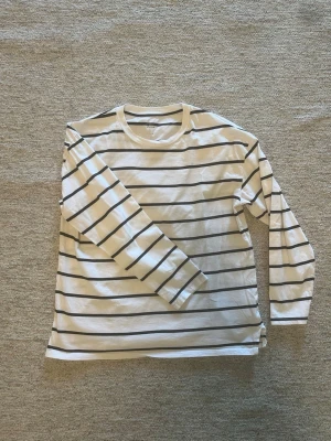 Vit långärmad randig topp - Klassisk vit långärmad topp med svarta horisontella ränder. Tillverkad i mjuk bomull med rund halsringning och relaxed fit. Perfekt att styla med jeans eller kjol för en enkel och clean look.