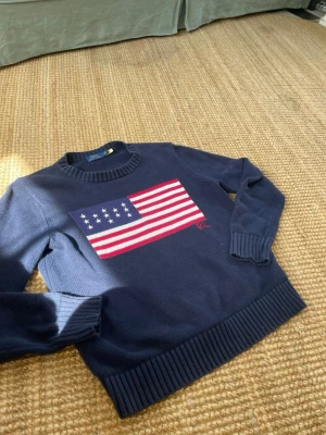Mörkblå stickad tröja Ralph Lauren - Mörkblå stickad tröja från Ralph Lauren med amerikansk flagga i rött, vitt och blått på bröstet. Tröjan har rund halsringning, ribbade muddar och RL-broderi under flaggan. Perfekt för dig som gillar klassisk och preppy stil.