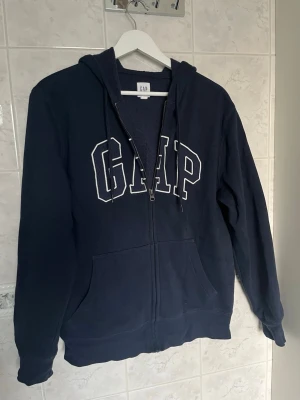 Mörkblå GAP zip hoodie med huva - Snygg mörkblå hoodie från GAP med dragkedja framtill och stor vit logga på bröstet. Tröjan har huva med snören och två stora fickor framtill. Nästan aldrig använd, köpt på GAP i London!💓
