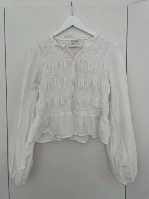 Vit blus med volang - Söt vit blus med volang nedtill. Blusen har rund halsringning, knäppning framtill och långa puffiga ärmar. Perfekt till jeans eller kjol för en trendig look.