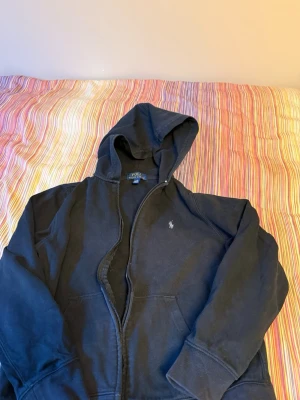 Svart hoodie från Polo Ralph Lauren - Säljer en svart hoodie från Polo Ralph Lauren med dragkedja och huva. Klassisk logga broderad på bröstet och två fickor framtill. Tillverkad i mjukt bomullsmaterial som är skönt att ha på sig. Perfekt för en avslappnad och stilren look.