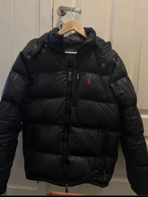 Svart pufferjacka från Polo Ralph Lauren - Säljer en svart pufferjacka från Polo Ralph Lauren med huva och dragkedja framtill. Jackan har två stora fickor med dragkedja och den klassiska röda loggan på bröstet. Perfekt för kalla vinterdagar och riktigt skön att bära.