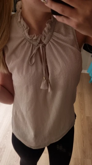 Beige ärmlös blus med volangkrage - Snygg beige blus med volangkrage och knytband framtill. Blusen är ärmlös och har en lätt, luftig känsla med rundad nederkant. Perfekt för en trendig och avslappnad look.