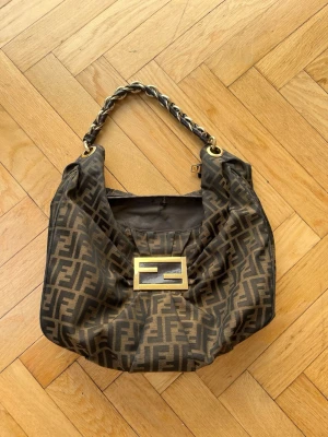 Fendi Mia monogram axelväska - Säljer min fina väska från Fendi med ikoniskt FF-monogram i tyg. Väskan har guldfärgad metallogo framtill och ett handtag i kedja och läder. Tillverkad i Italien och stängs med dragkedja. Har kommit väl till användning och har vissa bristningar (se bild) men inget som påverkar användningen. Är väldigt rymlig så perfekt vardagsväska!!
