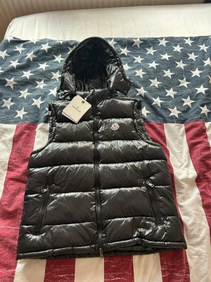 Svart dunväst från Moncler med huva - Snygg svart dunväst från Moncler med glansig finish och avtagbar huva. Västen har två dragkedjeförsedda fickor och klassisk Moncler-logga på bröstet. Perfekt för lager-på-lager och streetwear-stil.