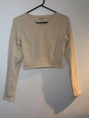 Beige ribbad långärmad crop top - Snygg beige ribbad crop top från Gina Tricot med långa ärmar och bred mudd i midjan. Tillverkad i stretchigt material som sitter skönt på kroppen. Perfekt till träning eller som ett stilrent basplagg.