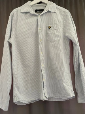 Ljusblå randig skjorta Lyle & Scott - Klassisk ljusblå skjorta från Lyle & Scott med tunna vita ränder. Skjortan har lång ärm, bröstficka med broderad logotyp och vita knappar framtill. Tillverkad i mjukt bomullstyg och har en avslappnad passform.
