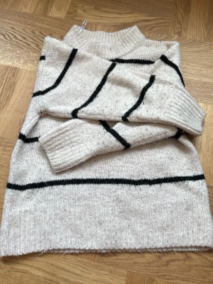 Beige stickad tröja Gina Tricot - Beige stickad tröja från Gina Tricot med svarta horisontella och vertikala ränder. Tröjan har hög krage och ribbade muddar vid ärmslut och nederkant. Perfekt för dig som gillar enkel och stilren design.